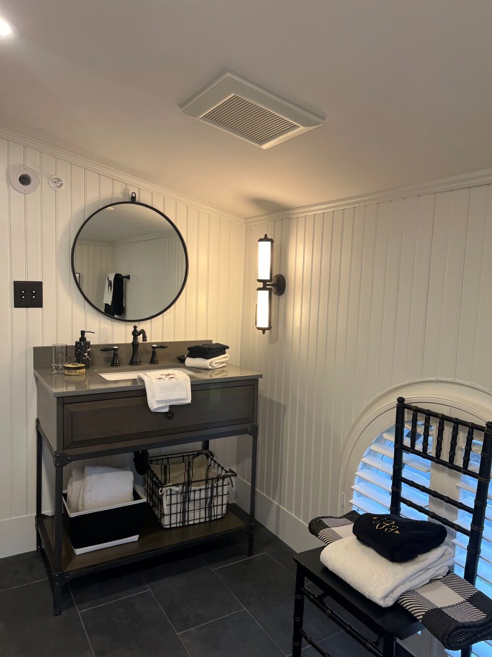 White paneled wall, dark tiled bathroom floor, black chair, white and black bath towels, dark bathroom sink, baskets