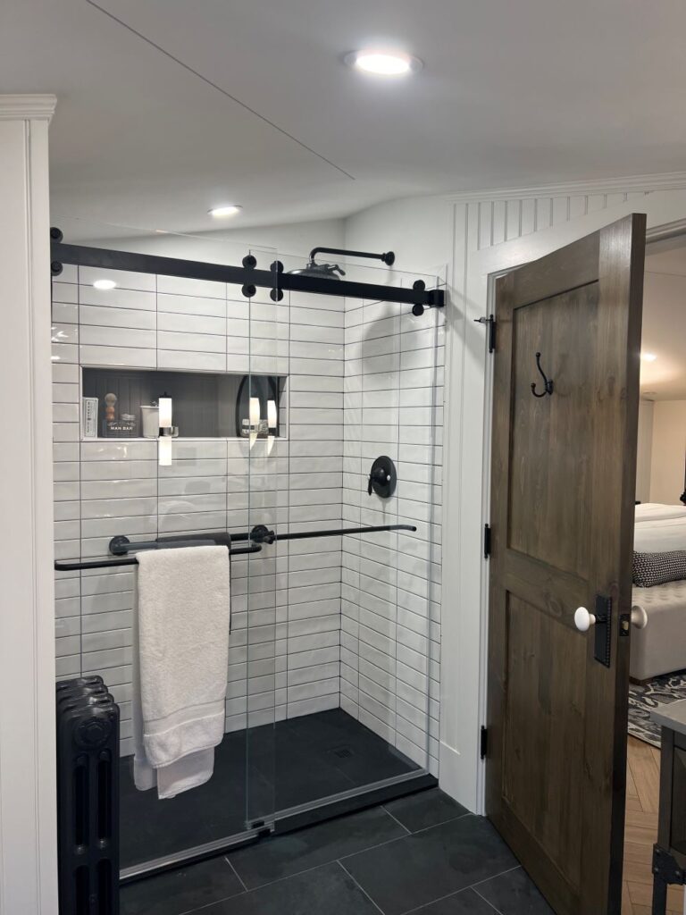 white shower wall subway tiles, dark bathroom tiles floors, white towel, black radiator, wooden door, black grab bars,
