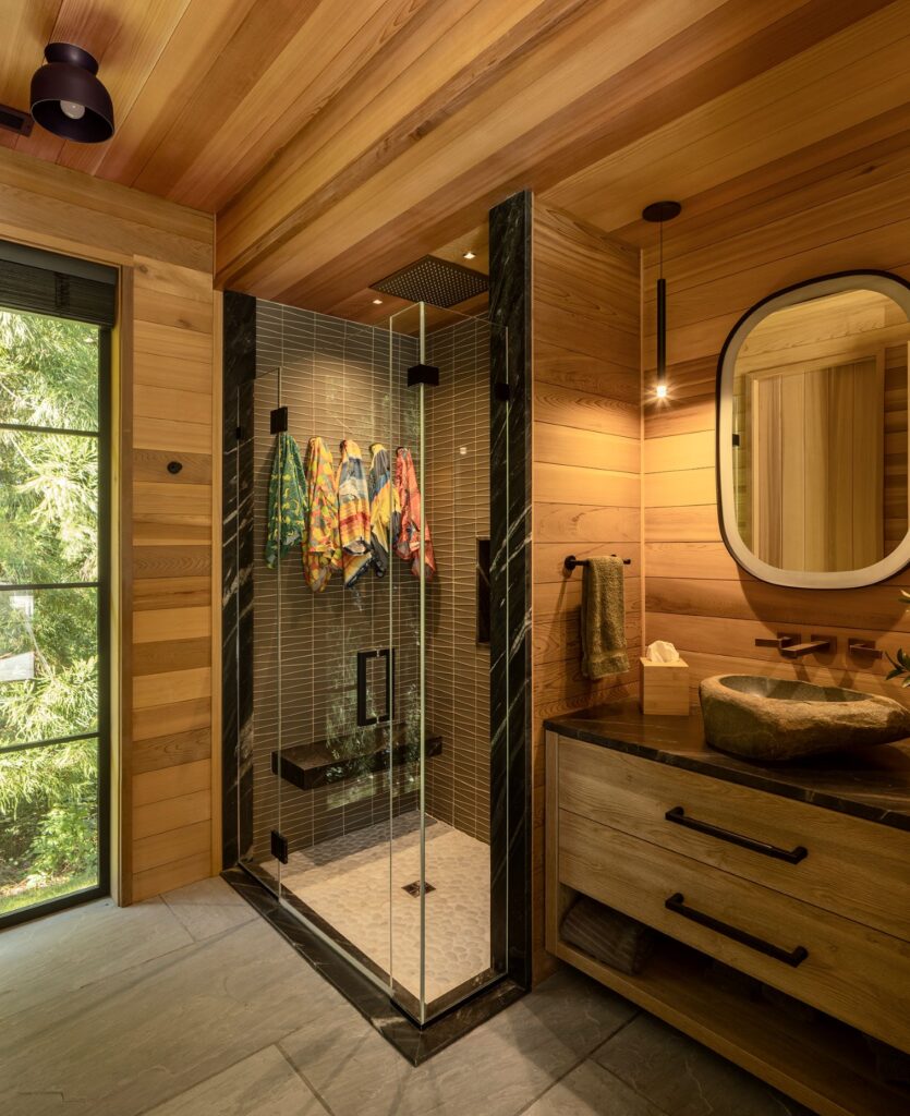 Wood-paneled bathroom with glass shower, colorful towels, stone sink, black countertop, and oval mirror, illuminated by natural light from a window.