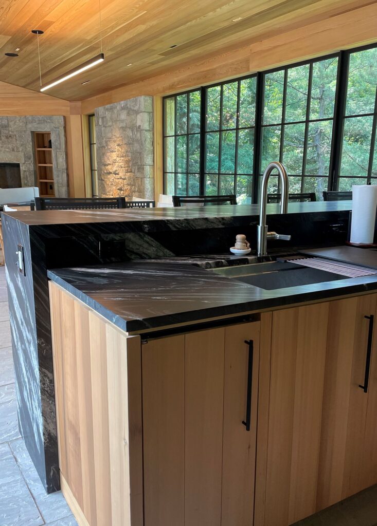 Wood paneled kitchen cabinets, black marble countertops, big windows, stone wall, chrome faucet, wood paneled ceiling